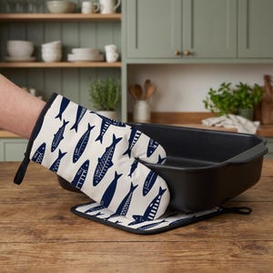 May include: An oven glove and a matching square pot holder featuring a repeating pattern of dark blue fish on a cream background. The oven glove is shown gripping the edge of a large, empty, rectangular black metal roasting pan on a wooden surface.