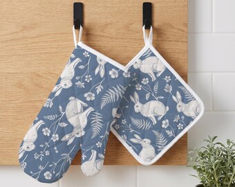 May include: A blue oven mitt and a square pot holder featuring a white line-art pattern of rabbits, ferns, and small flowers. Both items have white trim and white fabric loops for hanging, showcasing a consistent botanical and animal print design.