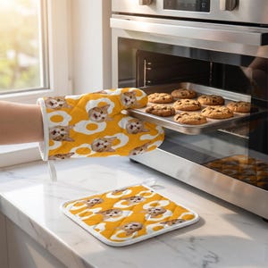 May include: A quilted oven glove and a matching square pot holder featuring a repeating pattern of orange cat faces centred within white fried egg shapes on a vibrant yellow background. The oven glove is shown in use near an open oven.