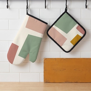 Op de afbeelding: Een ovenwant en een vierkante pannenlap met een geometrisch patroon van crème, saliegroene, oudroze en mosterdgele blokken. Beide items zijn voorzien van een zwarte stoffen rand en een zwarte ophanglus voor een strak en kleurrijk ontwerp.