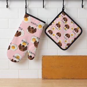 May include: A pink oven mitt and a square pot holder featuring a repeating pattern of brown dachshund dog faces, fried eggs, and the name Frankie printed in black text. Both items have black fabric trim and are hanging from black hooks.
