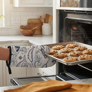 May include: An oven mitt and a matching square pot holder featuring a navy blue ink-style print of narwhals, orcas, and compass roses on a white background. The oven mitt has a black fabric trim and a hanging loop, while the pot holder is flat.