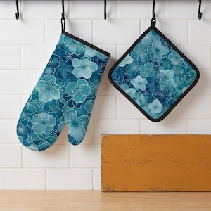 May include: An oven mitt and a square pot holder featuring a repeating floral pattern in shades of blue, teal, and white. Both items have black trim and are suspended from black hooks against a white tiled wall above a wooden surface.