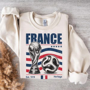 May include: A cream-colored crewneck sweatshirt featuring a graphic print with the word FRANCE in blue, a trophy illustration, a soccer ball, five blue stars, and red and blue wavy stripes. The text Est. 1919 and Heritage appear at the bottom.
