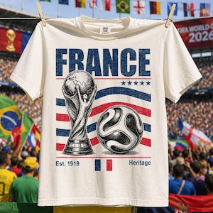 May include: An off-white t-shirt featuring the word FRANCE in blue, a graphic of a soccer trophy, a soccer ball, five blue stars, and the text Est. 1919 Heritage with a small French flag icon. The design uses blue and red horizontal stripes.