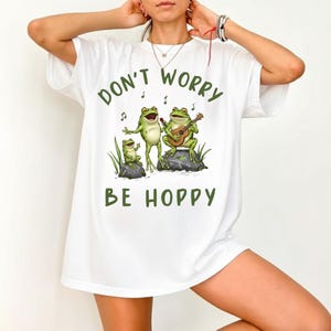 May include: White t-shirt featuring a green graphic of three frogs singing and playing a guitar on rocks. The text "DON'T WORRY BE HOPPY" is printed above and below the frogs in green. The shirt has short sleeves.