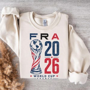 Op de afbeelding: Een cr&egrave;mekleurig sweatshirt met ronde hals en een grafische print met de tekst FRA 20 26 en WORLD CUP FRANCE. Het ontwerp bevat een gestileerde illustratie van een trofee met een wereldbol aan de bovenkant, in rode, witte en blauwe inkt.