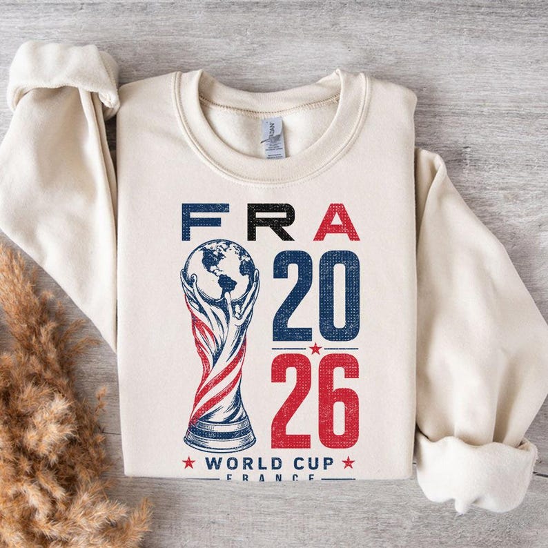 Op de afbeelding: Een cr&egrave;mekleurig sweatshirt met ronde hals en een grafische print met de tekst FRA 20 26 en WORLD CUP FRANCE. Het ontwerp bevat een gestileerde illustratie van een trofee met een wereldbol aan de bovenkant, in rode, witte en blauwe inkt.