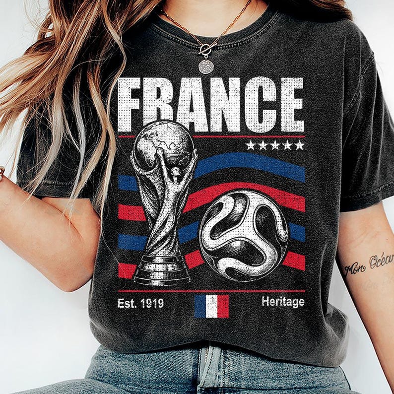 May include: A charcoal gray t-shirt featuring a graphic print with the word FRANCE in white, a soccer trophy, a soccer ball, five stars, red and blue curved lines, the text Est. 1919, a small French flag icon, and the word Heritage.