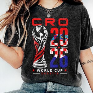 K&ouml;nnte beinhalten: Ein anthrazitfarbenes Kurzarm-T-Shirt mit einem Grafikdruck mit dem Text CRO 2026 WORLD CUP CROATIA. Das Design enth&auml;lt eine stilisierte Troph&auml;enillustration und die Zahl 2026, die mit dem rot-wei&szlig;en kroatischen Schachbrettmuster gef&uuml;llt ist.