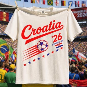 May include: An off-white short-sleeve t-shirt featuring the red text Croatia, the number 26, and the text Soccer Club. A graphic of a soccer ball with blue stars and red motion lines is displayed above a row of alternating red and blue stars.