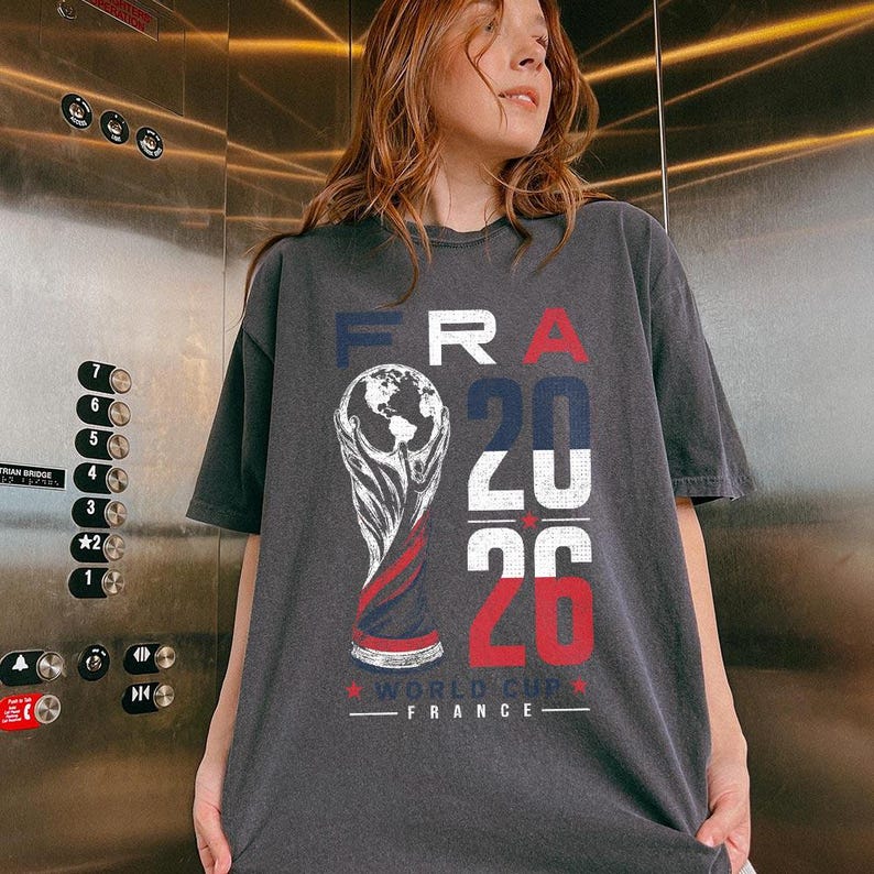 Op de afbeelding: Een antracietgrijs t-shirt met een grafische print van de Wereldbeker-trofee, de tekst FRA 2026 en de woorden WORLD CUP FRANCE. Het ontwerp bevat blauwe, witte en rode kleuren, met een verweerde textuur op de belettering en de trofee.