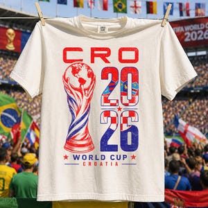K&ouml;nnte beinhalten: Ein cremefarbenes Kurzarm-T-Shirt mit einer Grafik des Weltpokals in Rot und Blau. Der Text lautet CRO 2026 WORLD CUP CROATIA, wobei die Zahl 2026 mit einem rot-wei&szlig;en Karomuster und blauen Akzenten gef&uuml;llt ist.