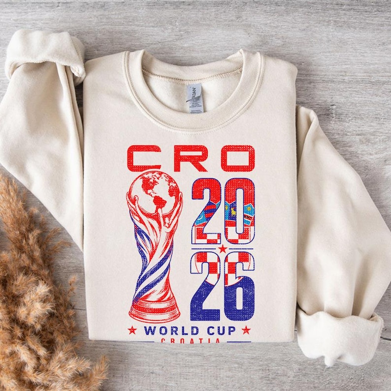 K&ouml;nnte beinhalten: Ein cremefarbenes Sweatshirt mit Rundhalsausschnitt und einem Grafikdruck mit dem Text CRO 2026 WORLD CUP CROATIA. Das Design enth&auml;lt eine stilisierte Pokalabbildung und die Ziffern 2026, die mit einem rot-wei&szlig;en Karomuster gef&uuml;llt sind.