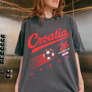 May include: A charcoal gray short-sleeve t-shirt featuring the red text Croatia, the number 26, and the words SOCCER CLUB. The graphic includes a red soccer ball with motion lines, a small Croatian flag, and a series of red and blue stars.
