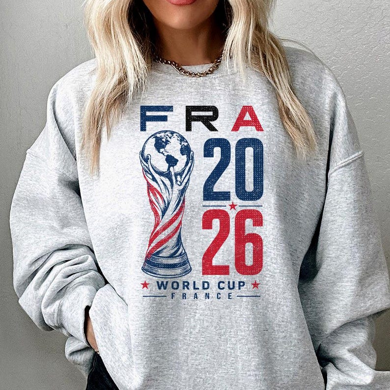 Op de afbeelding: Een lichtgrijs sweatshirt met ronde hals en een grafische print met de tekst FRA 20 26 en WORLD CUP FRANCE. Het ontwerp bevat een gestileerde illustratie van een trofee met een wereldbol aan de bovenkant in rood, wit en blauw met textuur.