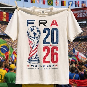 Op de afbeelding: Een cr&egrave;mekleurig t-shirt met korte mouwen en een grafische print met de tekst FRA 20 26 en WORLD CUP FRANCE. Het ontwerp bevat een gestileerde trofee-illustratie in blauw en rood, geaccentueerd met sterren, gecentreerd op de voorkant.