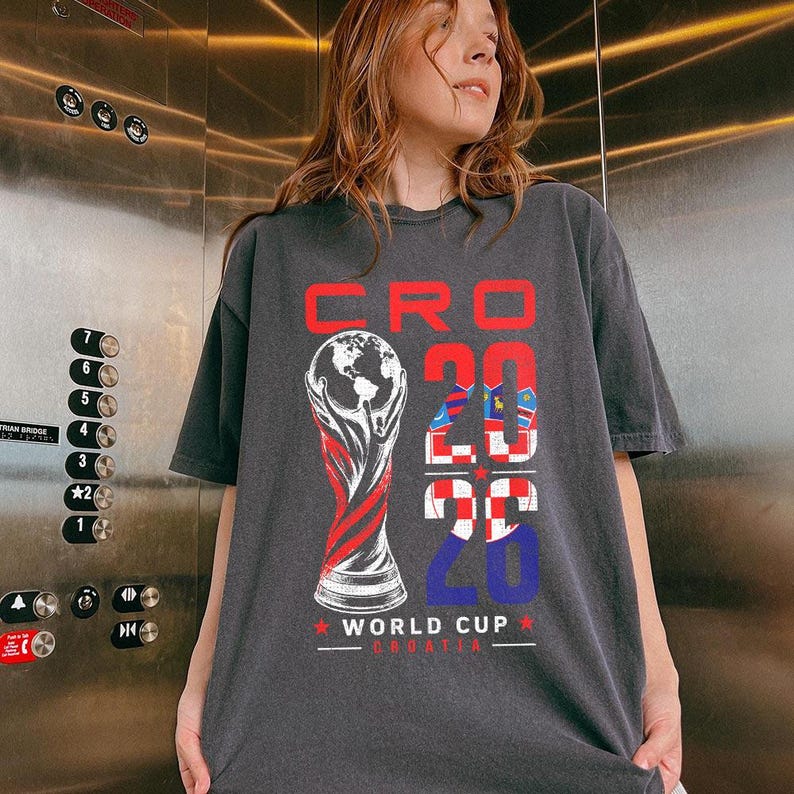 K&ouml;nnte beinhalten: Ein anthrazitfarbenes T-Shirt mit einem Grafikdruck und dem Text CRO 2026 WORLD CUP CROATIA. Das Design zeigt eine stilisierte Illustration eines Pokals mit einer Weltkugel sowie die Zahl 2026, gef&uuml;llt mit einem rot-wei&szlig;en Karomuster.