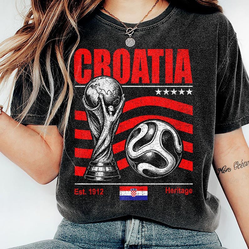 May include: A charcoal gray t-shirt featuring the word CROATIA in red letters above a graphic of a soccer trophy and a soccer ball. The design includes red horizontal stripes, five white stars, the text Est. 1912, Heritage, and a small Croatian flag.