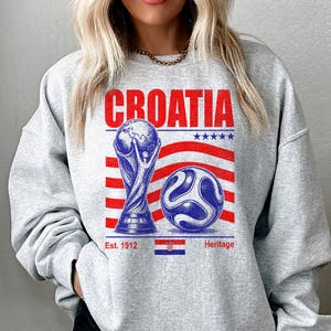 May include: A light gray crewneck sweatshirt featuring a graphic print with the word CROATIA in red, a gold-toned trophy, a blue and white soccer ball, red and white stripes, five blue stars, and the text Est. 1912 Heritage with a small flag.