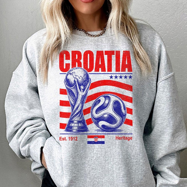 May include: A light gray crewneck sweatshirt featuring a graphic print with the word CROATIA in red, a gold-toned trophy, a blue and white soccer ball, red and white stripes, five blue stars, and the text Est. 1912 Heritage with a small flag.