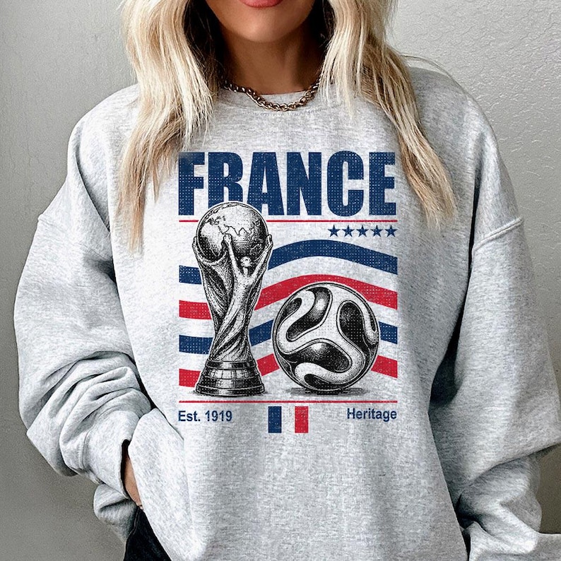 May include: A light gray crewneck sweatshirt featuring a graphic print with the text FRANCE, a trophy illustration, a soccer ball, five stars, and the text Est. 1919 Heritage. The design includes horizontal red and blue stripes and a small French flag.