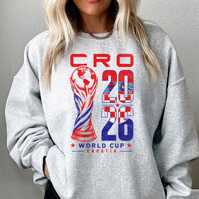 K&ouml;nnte beinhalten: Ein hellgraues Sweatshirt mit Rundhalsausschnitt und einem Grafikdruck mit dem Text CRO 2026 WORLD CUP CROATIA. Das Design enth&auml;lt eine stilisierte Troph&auml;enabbildung und die Zahl 2026, gef&uuml;llt mit dem rot-wei&szlig;en Karomuster der kroatischen Flagge.