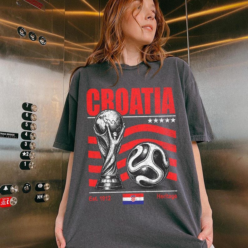May include: A charcoal gray t-shirt featuring a red graphic print that reads CROATIA. The design includes a soccer trophy, a soccer ball, five white stars, and the Croatian flag with the text Est. 1912 Heritage printed in red below the flag.