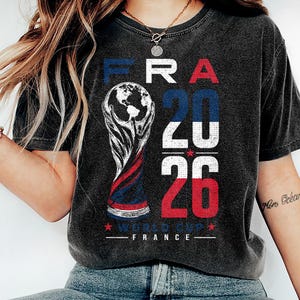 Op de afbeelding: Een antracietgrijs t-shirt met korte mouwen en een grafische print met de tekst FRA 2026 WORLD CUP FRANCE. Het ontwerp bevat een gestileerde trofee-illustratie en de cijfers 20 en 26 in blauw, wit en rood met een verweerde textuur.