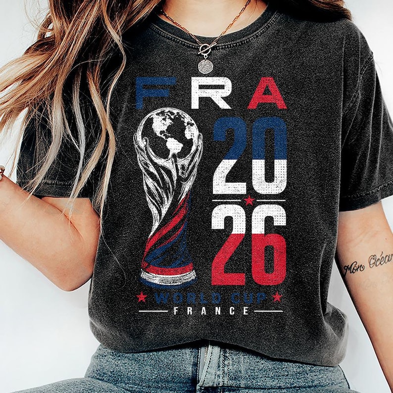 Op de afbeelding: Een antracietgrijs t-shirt met korte mouwen en een grafische print met de tekst FRA 2026 WORLD CUP FRANCE. Het ontwerp bevat een gestileerde trofee-illustratie en de cijfers 20 en 26 in blauw, wit en rood met een verweerde textuur.