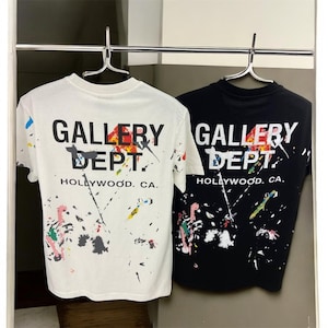 Könnte beinhalten: Zwei T-Shirts hängen an einem Kleiderbügel. Eines ist weiß, das andere schwarz. Beide haben den Text "GALLERY DEPT. HOLLYWOOD. CA." auf der Rückseite mit bunten Farbspritzern. Das weiße Shirt hat ein spärlicheres Spritzmuster.