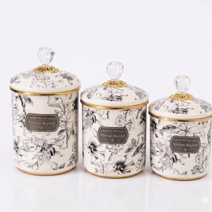 Peut inclure: Ensemble de trois bocaux de cuisine en céramique avec couvercles. Les bocaux blancs présentent un motif floral noir et gris, des bordures dorées et des boutons en cristal. Chaque bocal porte l'inscription "Kitchen Beyond".