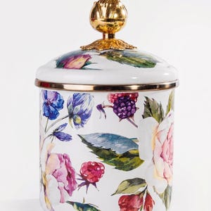 May include: White ceramic jar with a lid, decorated with a floral pattern of pink, purple, and blue flowers, and green leaves. The lid is topped with a gold-coloured finial. The jar has a gold rim.