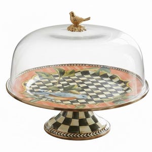 May include: A decorative cake stand with a clear glass dome. The plate features a black and white checkered pattern with a bird and leaf design. The base is also checkered, and a gold-colored bird tops the dome.