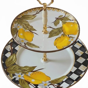 Puede incluir: Bandeja de dos niveles con asa y detalles dorados. El nivel superior presenta un fondo blanco con limones amarillos y hojas verdes. El nivel inferior tiene un patr&oacute;n de cuadros blancos y negros con detalles de lim&oacute;n y hojas.