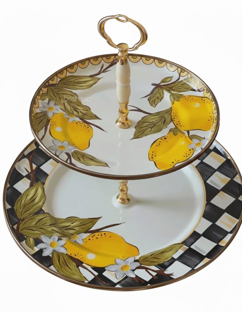 Puede incluir: Bandeja de dos niveles con asa y detalles dorados. El nivel superior presenta un fondo blanco con limones amarillos y hojas verdes. El nivel inferior tiene un patr&oacute;n de cuadros blancos y negros con detalles de lim&oacute;n y hojas. Decorativo.
