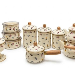 Puede incluir: Un conjunto de utensilios de cocina de esmalte color crema con un delicado patrón floral de pequeñas flores rojas, azules y amarillas. El set incluye un cuenco con pedestal, tres recipientes redondos apilados, tres botes, un cazo y una tetera doble.