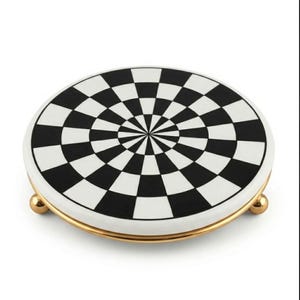 Puede incluir: Un soporte de exhibición circular de cerámica con un patrón de tablero de ajedrez en blanco y negro que irradia desde el centro en un diseño geométrico. La parte superior plana está sostenida por un borde de metal dorado pulido y tres pequeñas patas doradas redondeadas.