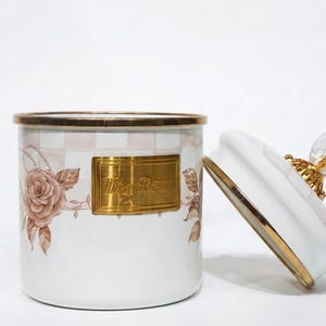 Può includere: Vaso in ceramica bianca con coperchio, con bordo e dettagli dorati. Il vaso è decorato con un motivo floreale e una placca dorata con la scritta "Miss Betty". Il coperchio ha un pomello di cristallo.