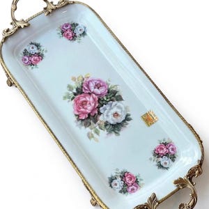 Puede incluir: Una bandeja rectangular con fondo azul claro y diseño floral de rosas rosas y blancas. La bandeja tiene asas y ribetes dorados. La bandeja mide aproximadamente 40 cm de largo.