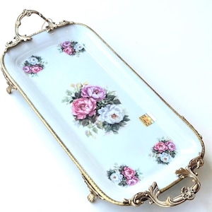 Puede incluir: Una bandeja de servicio rectangular de porcelana blanca con ramos de rosas rosas y blancas. La bandeja está enmarcada por un borde metálico dorado ornamentado con asas decorativas y cuatro pequeñas patas en la base.