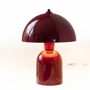 May include: A glossy, claret-coloured table lamp featuring a rounded base and a dome-shaped shade. The lamp has a small, round finial atop the shade. The design is modern and minimalist, with a focus on smooth, reflective surfaces.