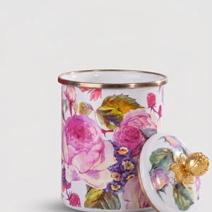 Può includere: Un barattolo di smalto bianco con un design floreale con rose rosa, fiori viola e foglie verdi. Il bordo è rifinito con oro. Il coperchio, anch'esso bianco, ha una maniglia a forma di rosa dorata.