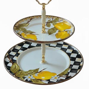Puede incluir: Bandeja de servir de dos niveles con asa dorada y soporte color crema. El nivel superior presenta un dise&ntilde;o de lim&oacute;n y hojas, mientras que el nivel inferior tiene un borde a cuadros en blanco y negro con un dise&ntilde;o de lim&oacute;n y flores.