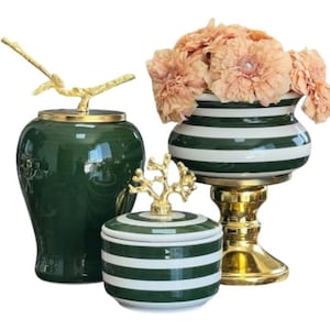 May include: Three decorative ceramic pieces: a dark green jar with a gold bird-shaped lid, a small striped container with a gold tree-shaped lid, and a striped vase with peach-colored flowers, set on a gold stand.