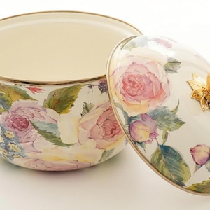 Puede incluir: Una olla de esmalte con estampado floral y tapa a juego. La olla presenta una base de color crema adornada con rosas rosas y moradas y hojas verdes. La tapa tiene un asa dorada en forma de rosa. El borde y las asas también son dorados.