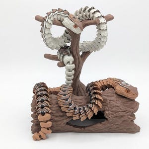 May include: An articulated snake figure with a white and brown segmented body coiled around a brown tree branch stand. A second articulated snake figure with a brown and black patterned body rests on a brown log base with a hollow opening.