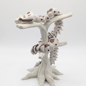 Puede incluir: Una figura de serpiente articulada blanca con manchas marrones y ojos naranjas, enroscada alrededor de un soporte con forma de árbol blanco. La serpiente presenta partes del cuerpo segmentadas que permiten el movimiento sobre la base texturizada.