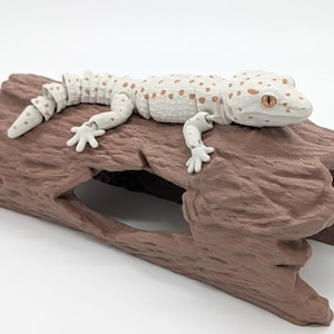 May include: An articulated white gecko figurine with brown spots and orange eyes resting on a brown, textured, hollow log structure. The gecko features segmented body parts that allow for movement, positioned horizontally across the top of the log.
