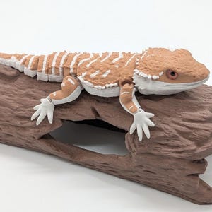 May include: A tan and white articulated bearded dragon figurine with a textured, scaly appearance, resting on top of a brown, hollow, log-shaped structure. The lizard features visible joints at the limbs and tail, with a detailed head and eyes.